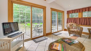 A spacious room with a large sliding glass door leading to an outdoor patio, a comfortable chair, a small table with a book, and a laptop on a bench. - Walk to Water: Outdoor Gem w/ Bunkhouse in Madison (Madison)