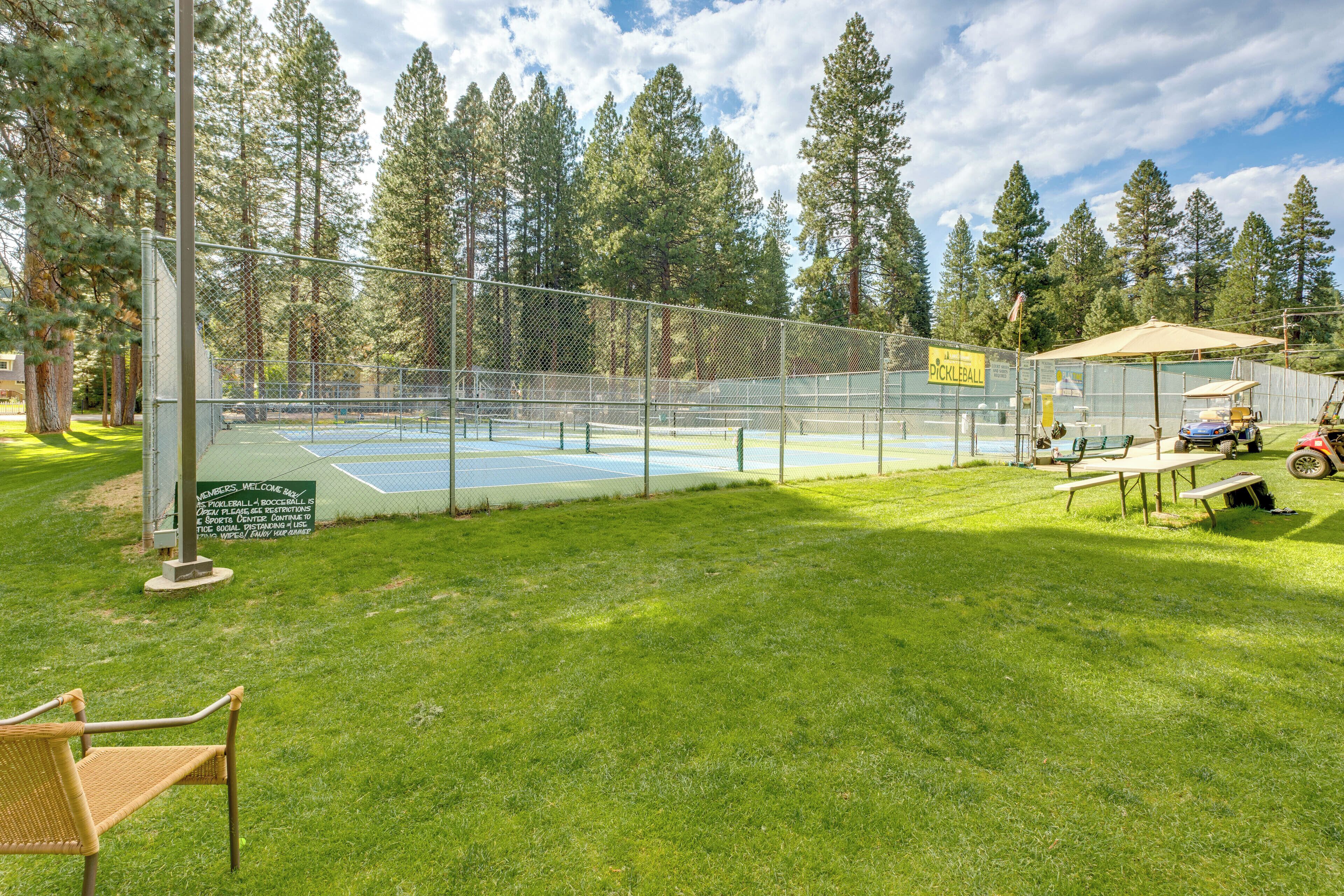 Una cancha de tenis rodeada por una cerca de malla metálica, con mesas de picnic y un carrito de golf cerca.