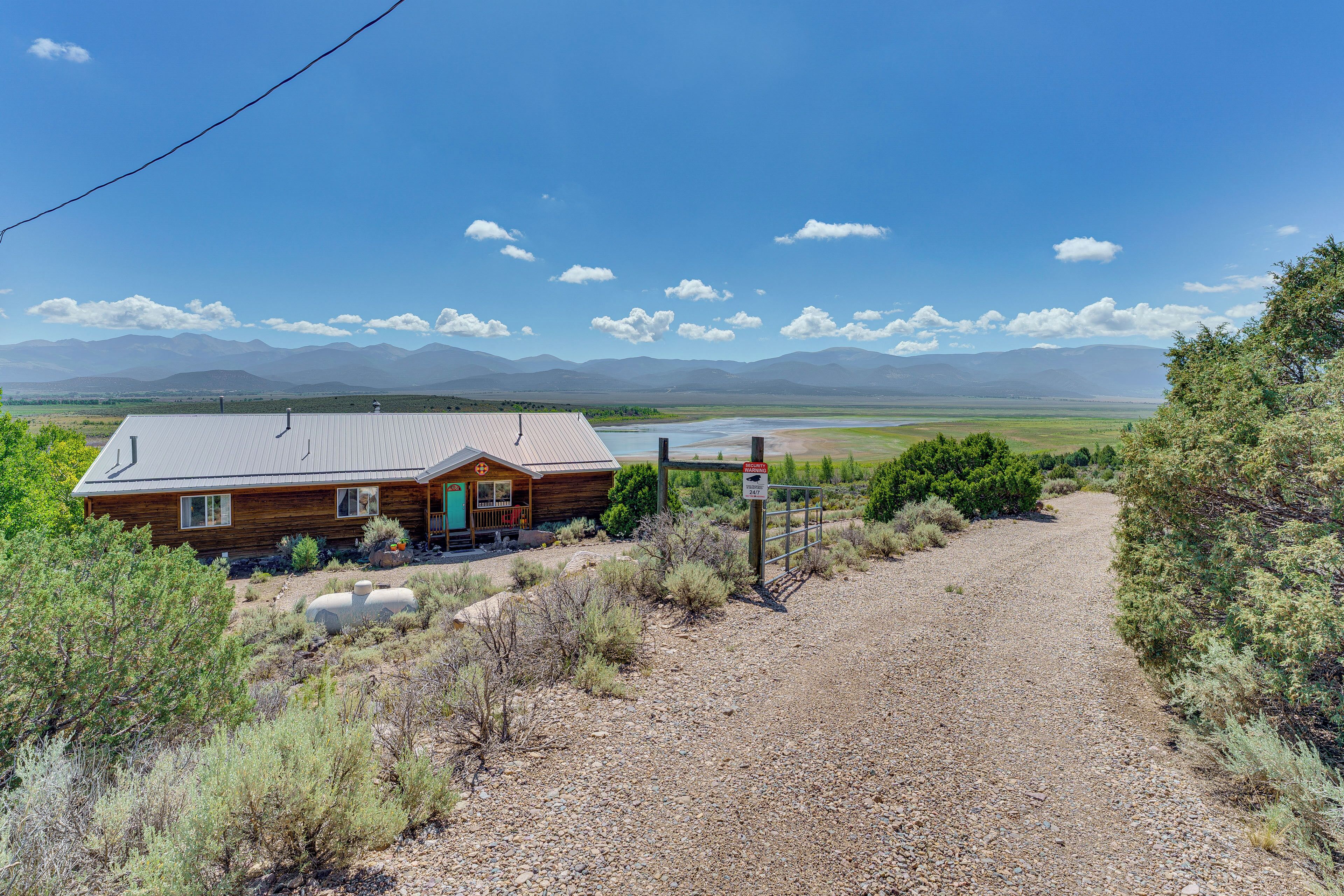 A rustic cabin with a gravel driveway, surrounded by dry vegetation and mountains in the background.
