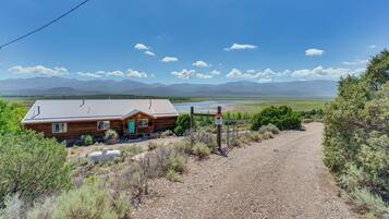 A rustic cabin with a gravel driveway, surrounded by dry vegetation and mountains in the background.