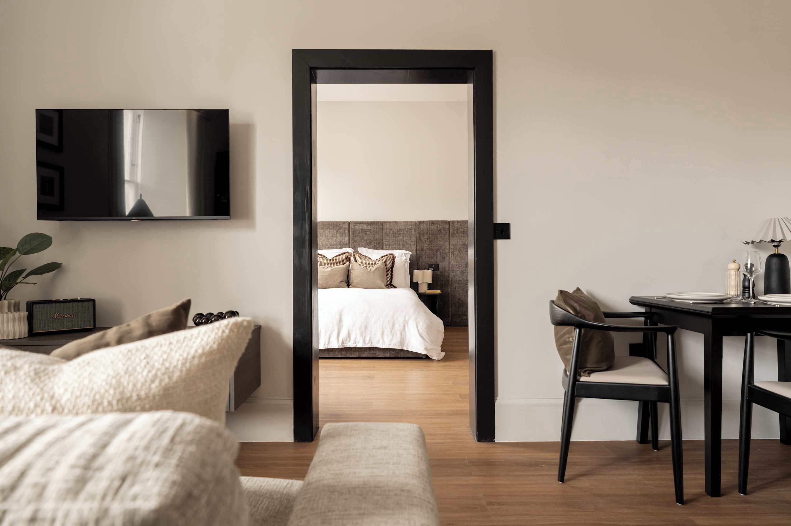 A modern hotel room with a large bed, a TV mounted on the wall, a dining area with a table and chairs, and a wooden floor.