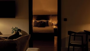 Ein schummrig beleuchteter Raum mit einem Bett, einem Nachttisch mit Lampe, einem Sessel und einem an der Wand befestigten Fernseher.