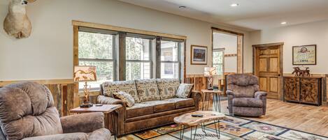 A cozy living room with a brown leather sofa, two armchairs, a coffee table, and a large rug with animal prints. A deer head is mounted on the wall, and there are large windows with wooden frames.