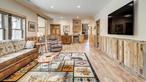 A spacious living room with a sofa, armchair, and coffee table, leading to a kitchen area with wooden cabinets and a TV mounted on the wall. - 3 MI to Lake: Pet Friendly Wabeno Cabin! (Wabeno)