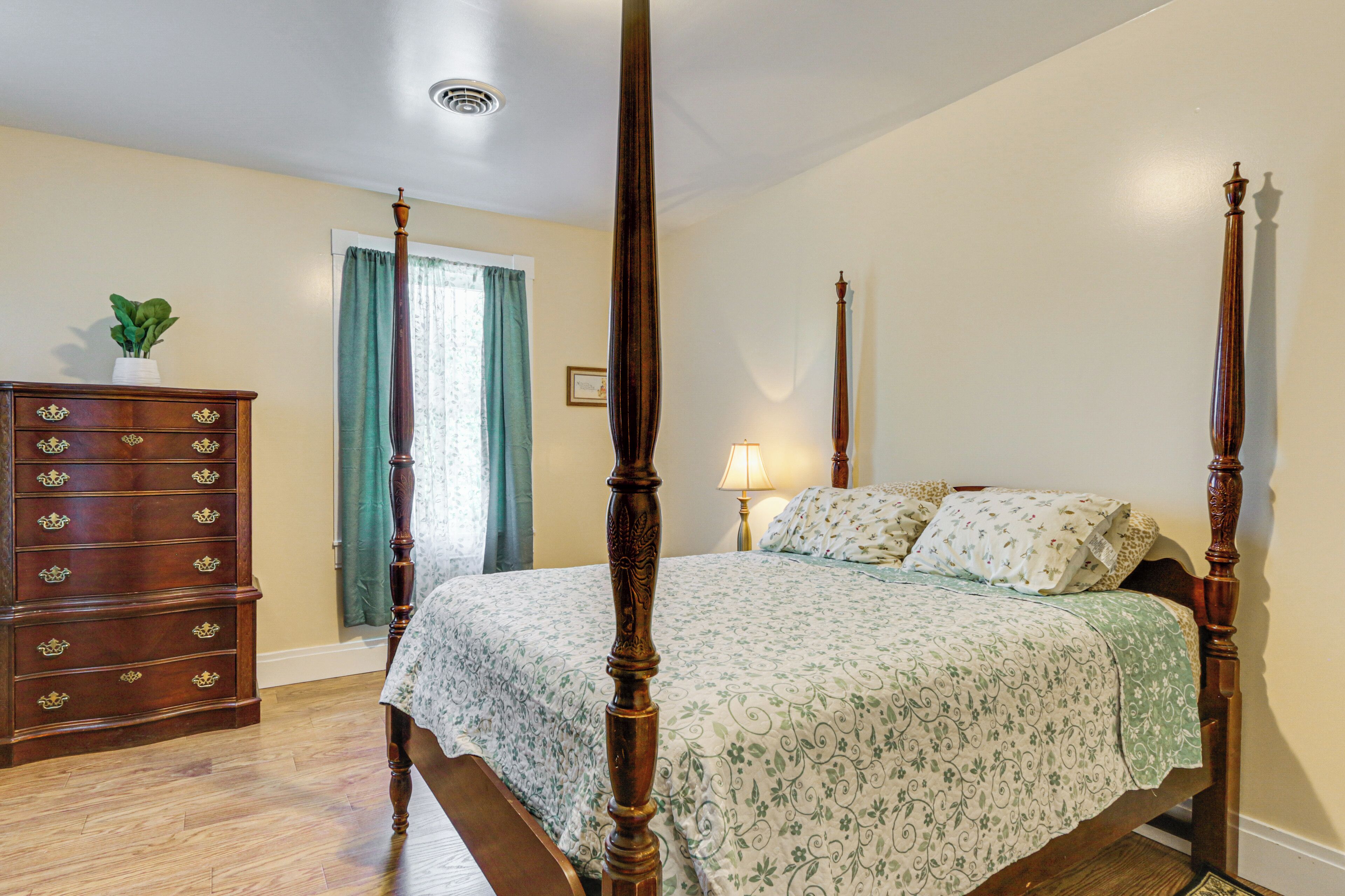 A four-poster bed with a floral bedspread, a wooden dresser with multiple drawers, and a window with curtains.