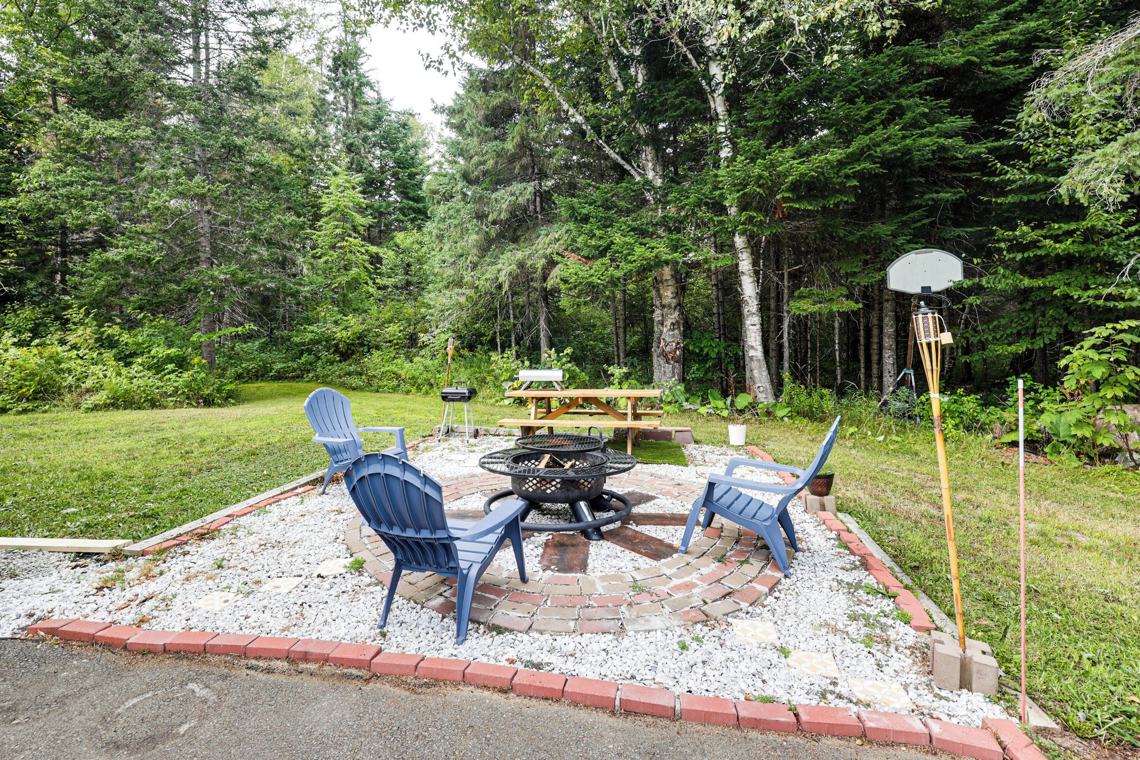 An outdoor patio area with a fire pit, two blue Adirondack chairs, and a picnic table surrounded by a brick border.