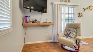 A living room with a mounted TV, a rocking chair, a window with blinds, and a shelf with books and decorative items. - 3 Mi to Icw: Coastal Carolina Home w/ Pool Access (Hampstead)