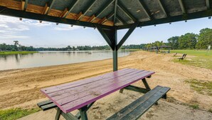 A covered picnic area with a bench and picnic table, overlooking a lake with a sandy shore and trees in the background. - Relax By the Lake: Quiet East Texas Retreat (Village Mills)
