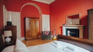 Une chambre d’hôtel avec un grand lit, une armoire en bois, un fauteuil gris, une guitare et une cheminée.