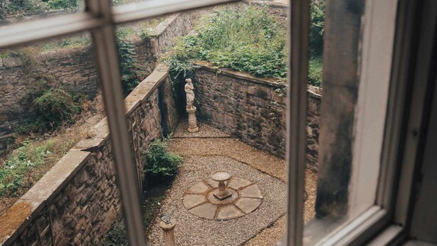 Une vue à travers une fenêtre donnant sur une cour avec des murs en pierre, une statue et un motif circulaire en pierre sur le sol.