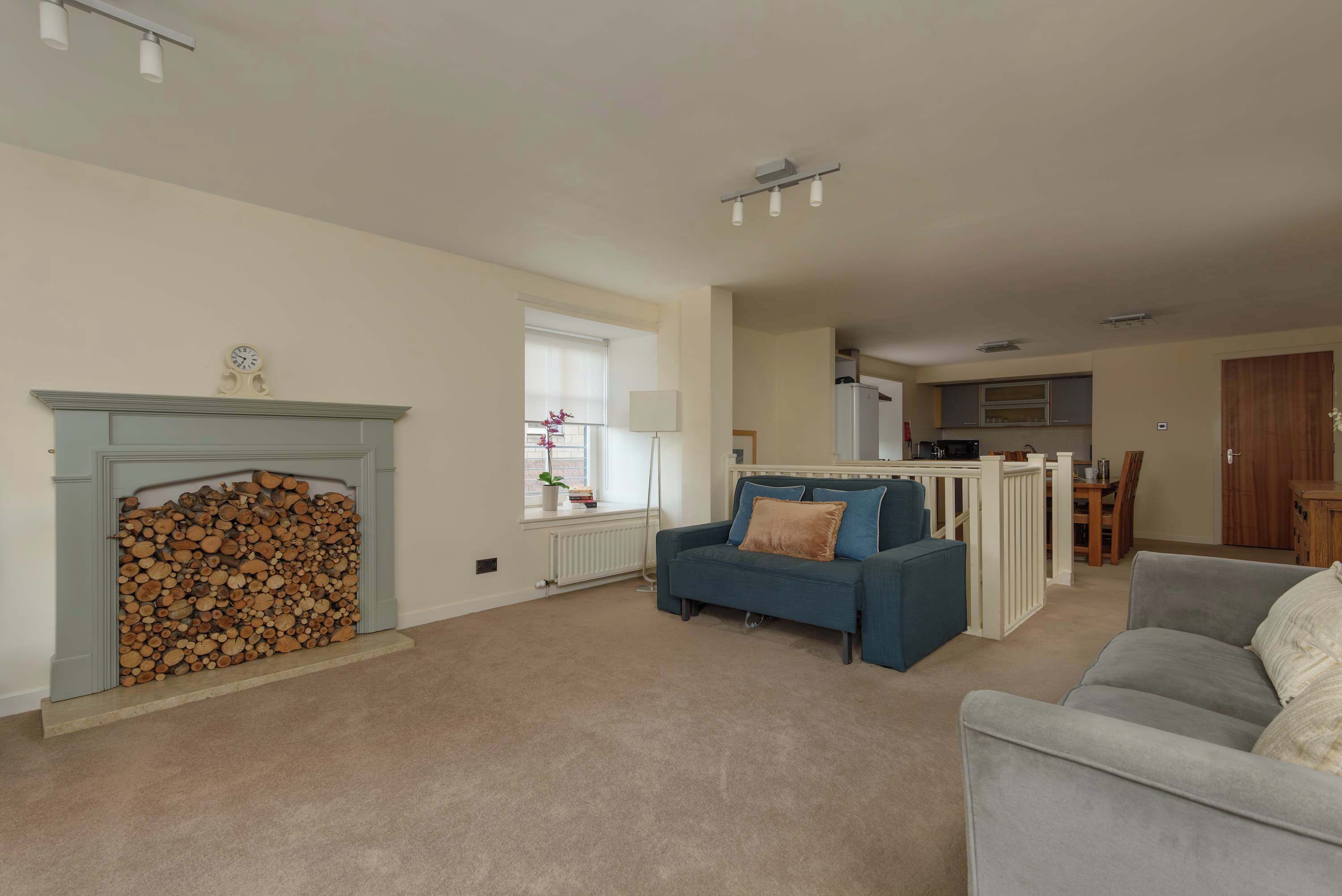A living room with a fireplace, a blue sofa, a grey sofa, and a kitchen area in the background.