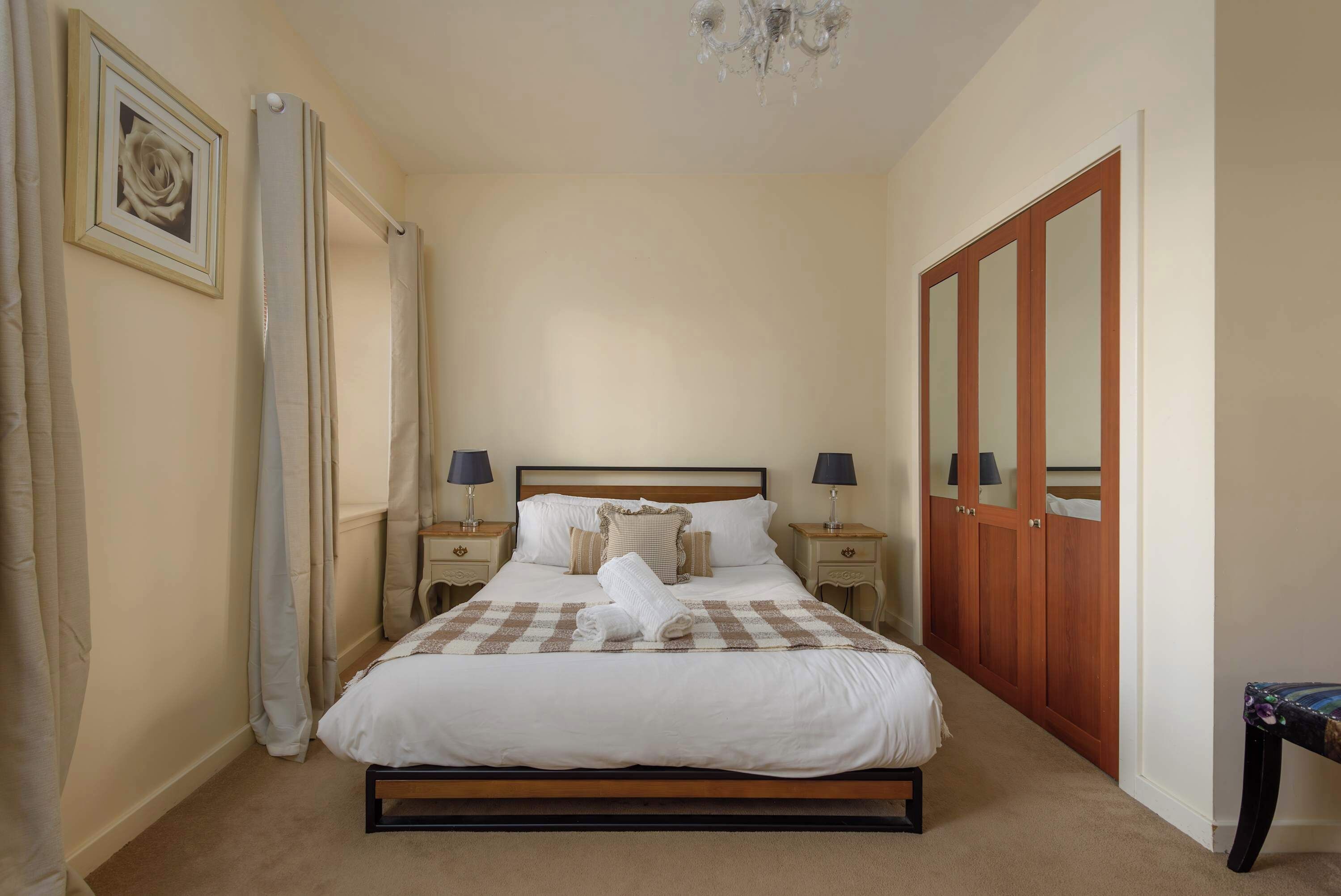 A neatly made bed with a checkered bedspread, two bedside lamps, a wooden wardrobe, and a framed picture on the wall.