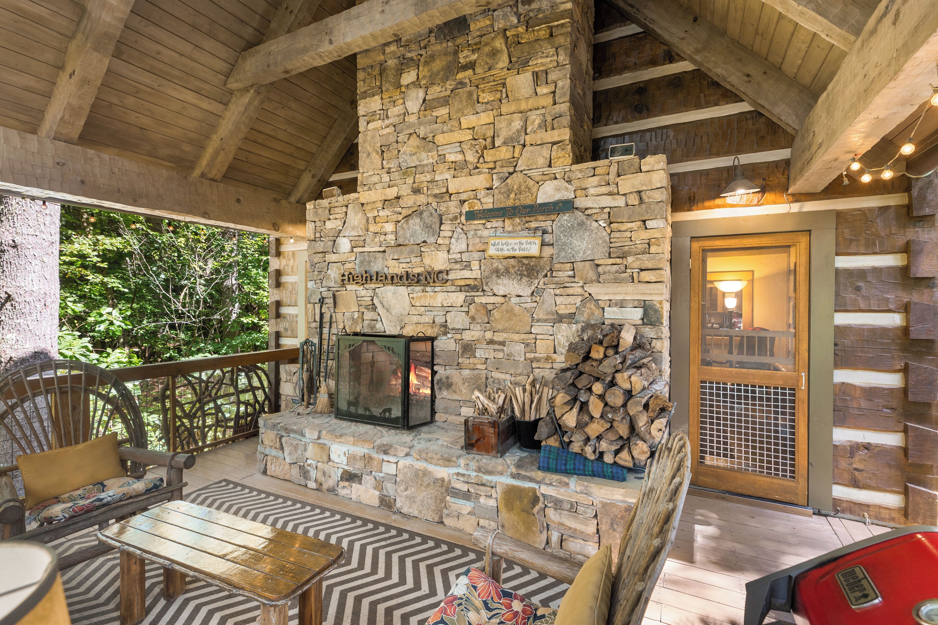 A stone fireplace with a log storage area, wooden furniture, and a view of the outdoors.