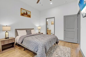 Ein Schlafzimmer mit Bett, Nachttischen, einer Lampe, einem Gemälde, einer Tür und einem Fernseher.