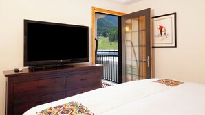 Een hotelkamer met een grote flatscreen-tv op een houten dressoir, een bed met een kleurrijke deken, een deur naar een balkon met uitzicht op de bergen, en een ingelijst foto aan de muur.