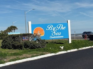 Une enseigne pour le complexe résidentiel « By The Sea Condominiums », représentant une scène de plage, un lampadaire et une route avec une voiture.