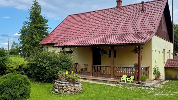 Une maison de plain-pied avec un toit recouvert de tuiles rouges, une véranda avec des chaises et un jardinière en pierre ornée de plantes.