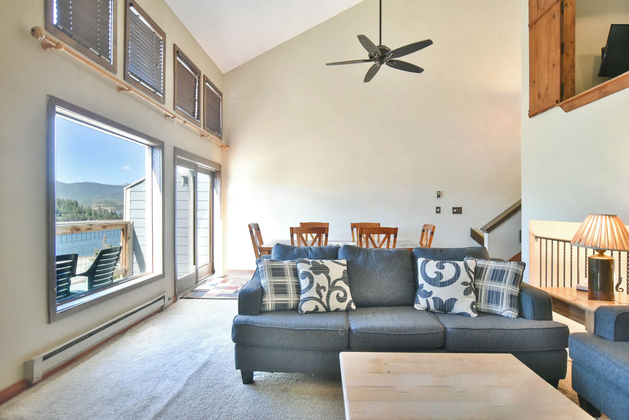 A spacious living room with a gray sectional sofa, a wooden coffee table, and a ceiling fan. Large windows offer a view of the outdoors.