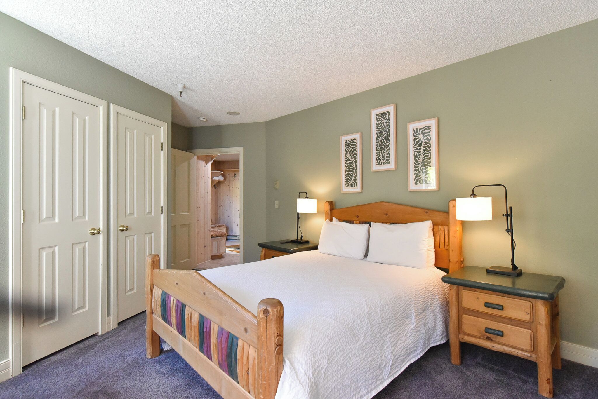 Une chambre à coucher avec un lit en bois, une literie blanche, deux tables de chevet avec des lampes et une porte donnant sur une salle de bain.