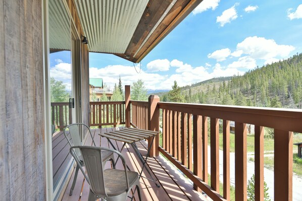 A wooden deck with chairs and a table, overlooking a forested mountain area.