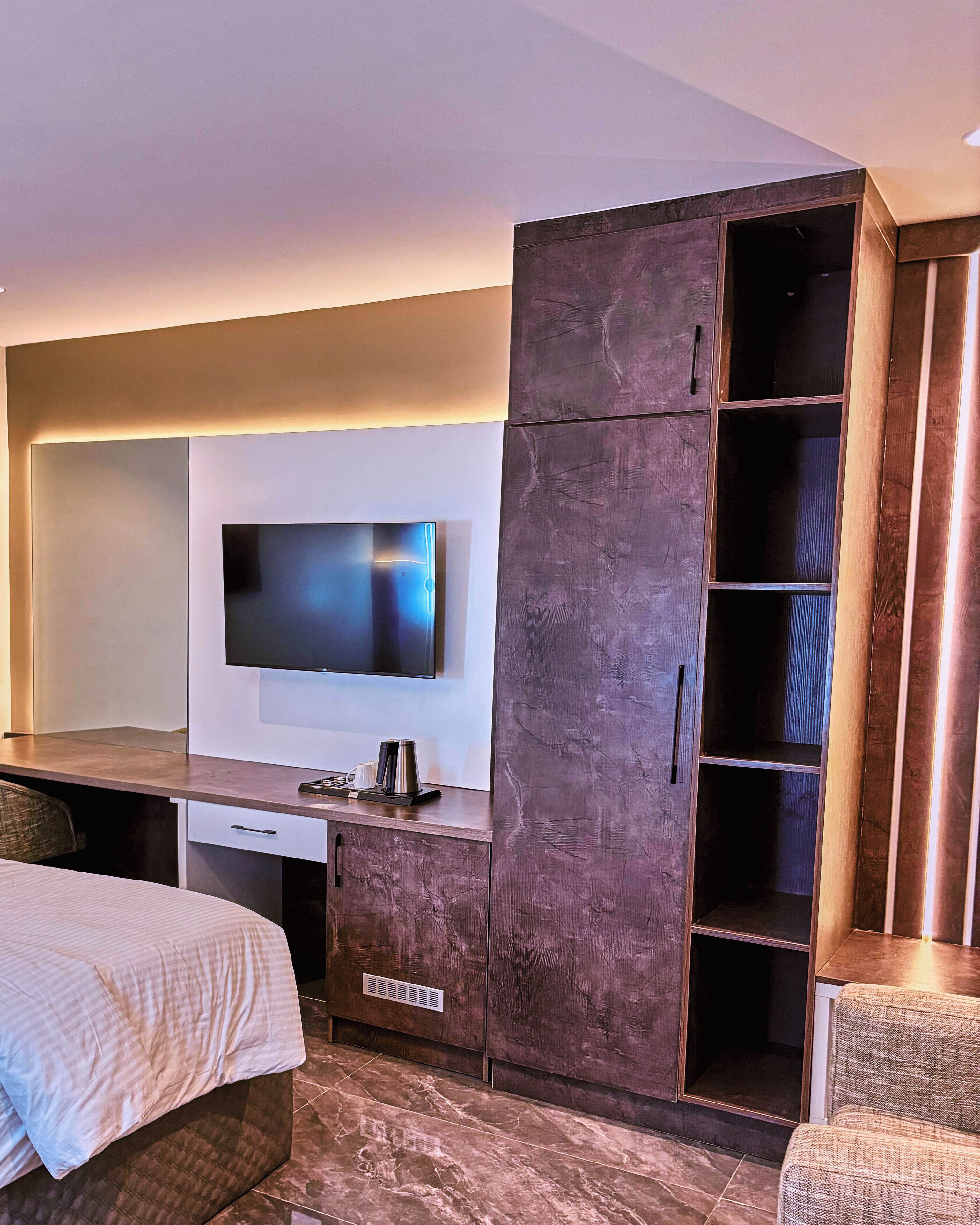 A modern hotel room with a flat-screen TV mounted on the wall, a wooden wardrobe with multiple shelves, a desk with a coffee maker, and a bed with a patterned bedspread.