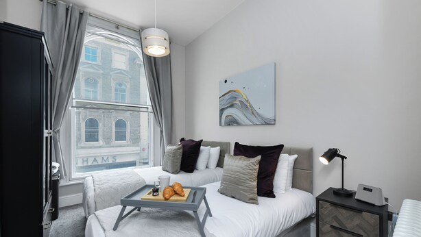 A hotel room with a bed, bedside table, lamp, and a view of a building through the window. - Market Street Apartment 3A Flat 1 (Nottingham)