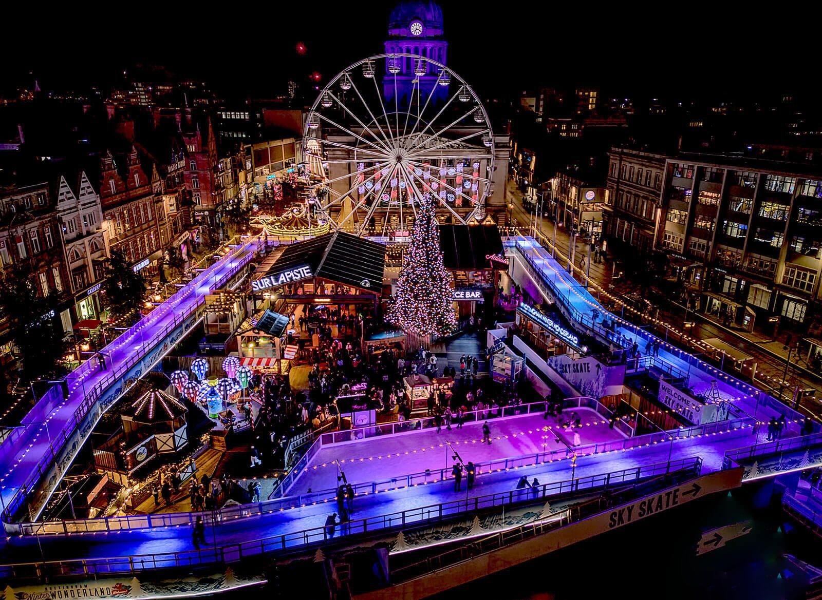A bustling nighttime cityscape with a prominent Ferris wheel, a large illuminated Christmas tree, and a skating rink.