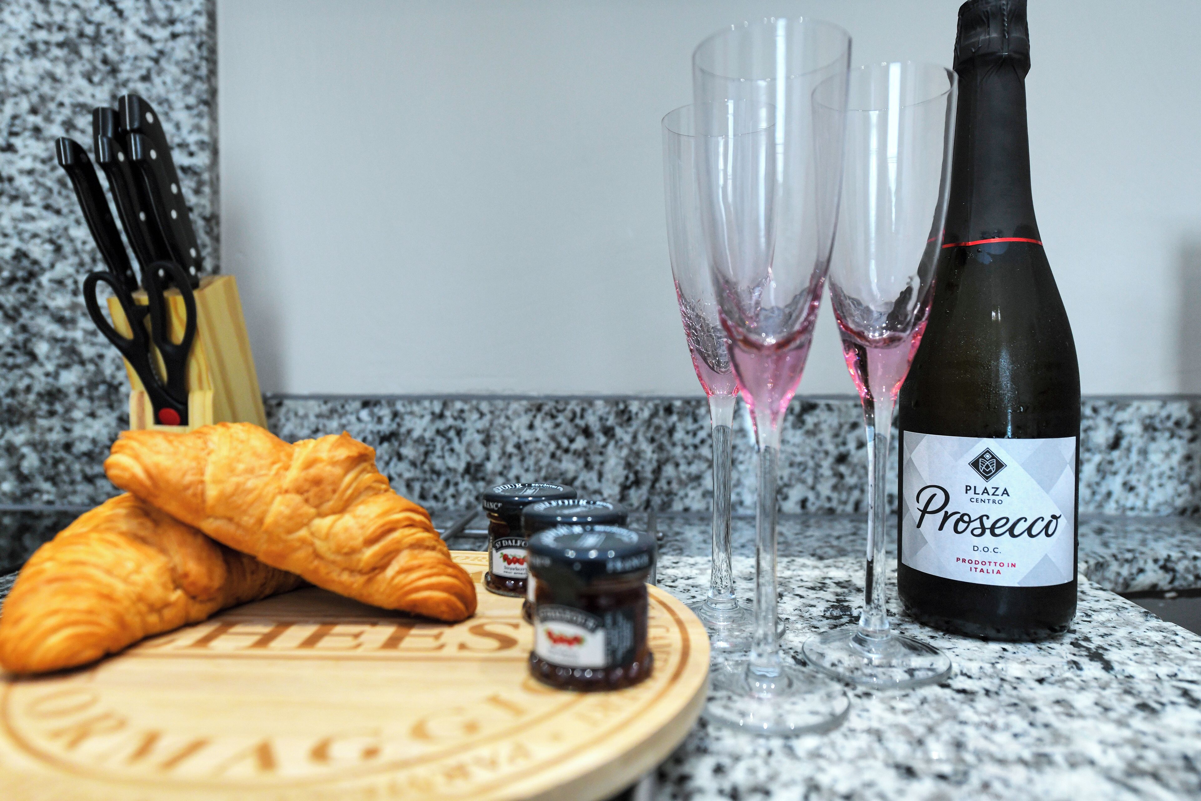Una encimera de cocina con una botella de Prosecco, dos vasos, un croissant y una tabla de cortar con la palabra 'FIEVES'.