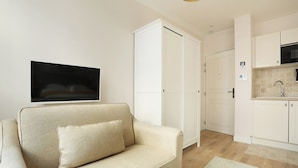 Ein Wohnzimmer mit beiger Couch, einem am Wand montierten Flachbildfernseher und einer Küchenzeile mit weißen Schränken und einer Mikrowelle.