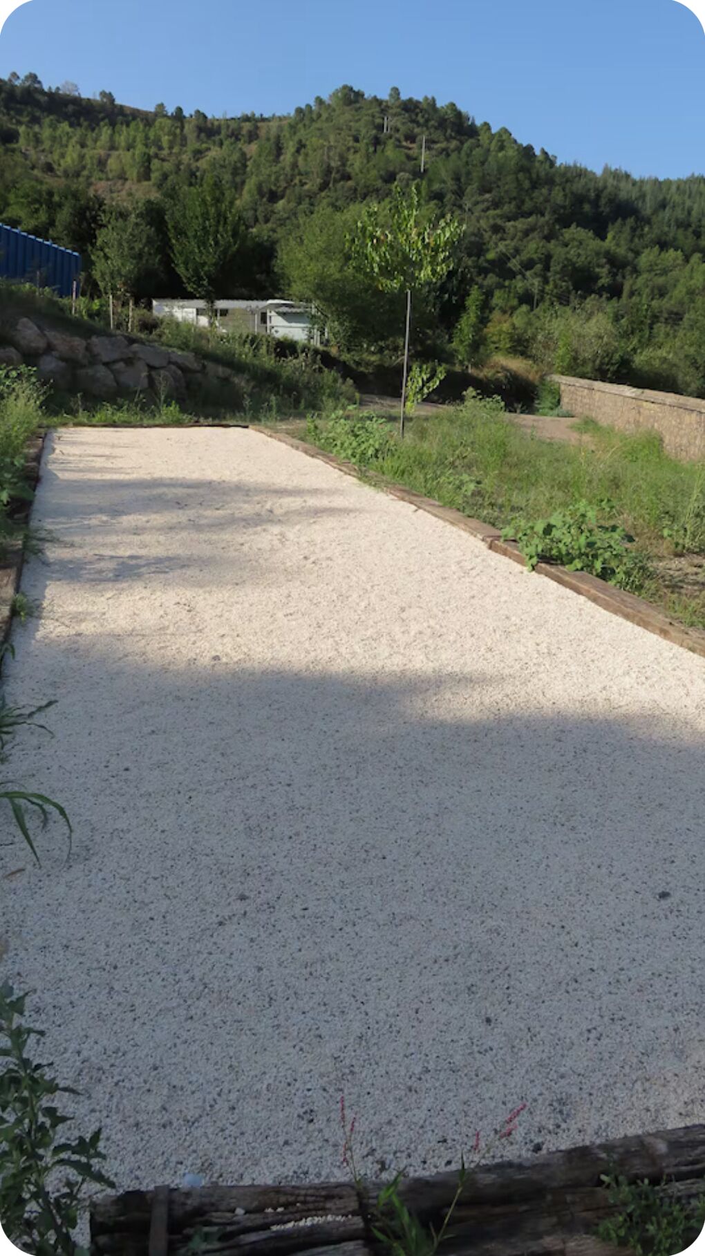 A gravel path leading through a rural area with a stone wall on one side and a forested hill in the background.