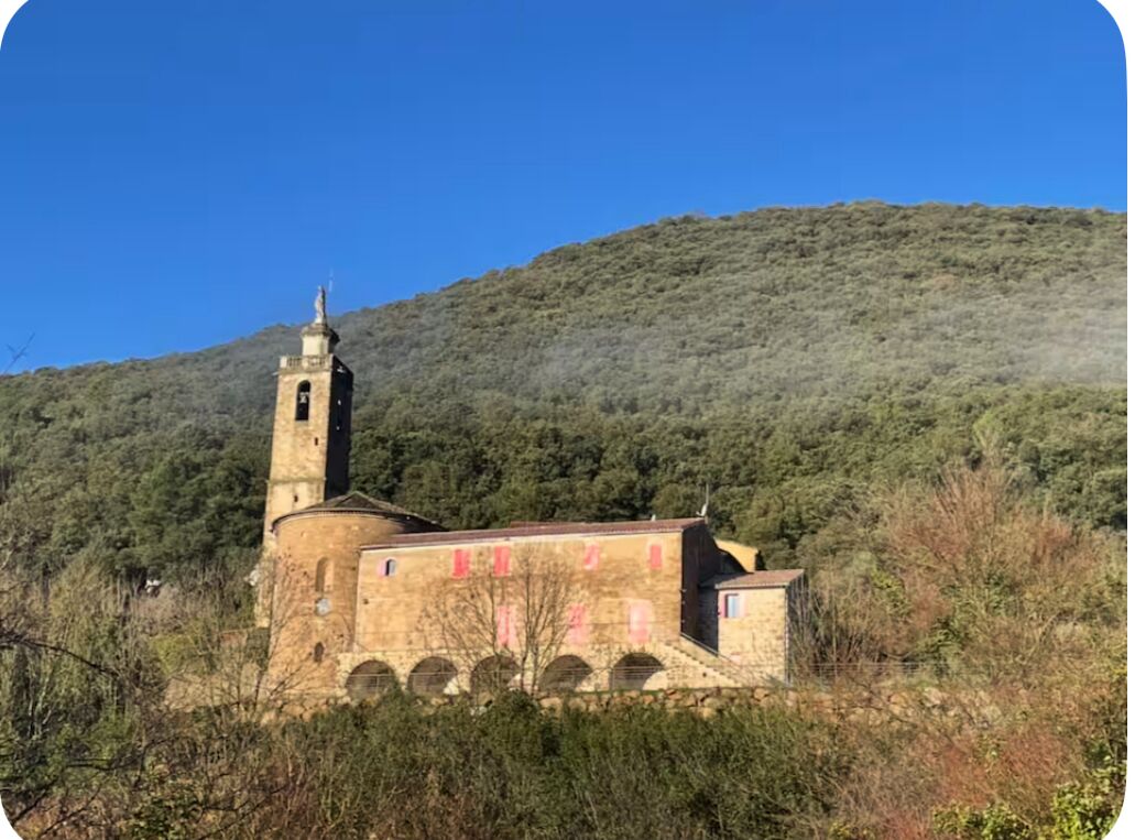 A stone church with a bell tower, surrounded by dense vegetation and hills.