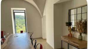 Una zona de menjador amb una taula de fusta llarga, caires i vista a un paisatge de muntanyes per una porta.