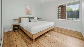 Un dormitorio con cama de madera, mesita de noche, lámpara de pie, ventilador de techo y ventana con vista a un muro de ladrillo y follaje.