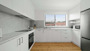 Una cocina moderna con armarios blancos, suelo de madera, un ventanal con vistas a edificios y un refrigerador de acero inoxidable.