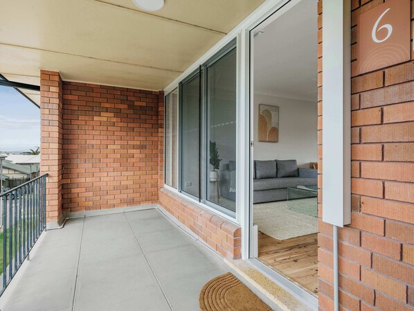 A balcony with a brick wall, a sliding glass door leading to a living room with a sofa and a rug. - Prickly Pear Stays in Merewether - Ranclaud St, 1-bedroom (Merewether)