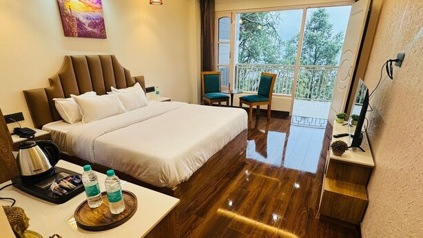 Habitación de hotel con una cama grande, un escritorio con hervidor, una botella de agua y una estatua de Buda. Tiene un balcón con sillas y vistas a árboles.