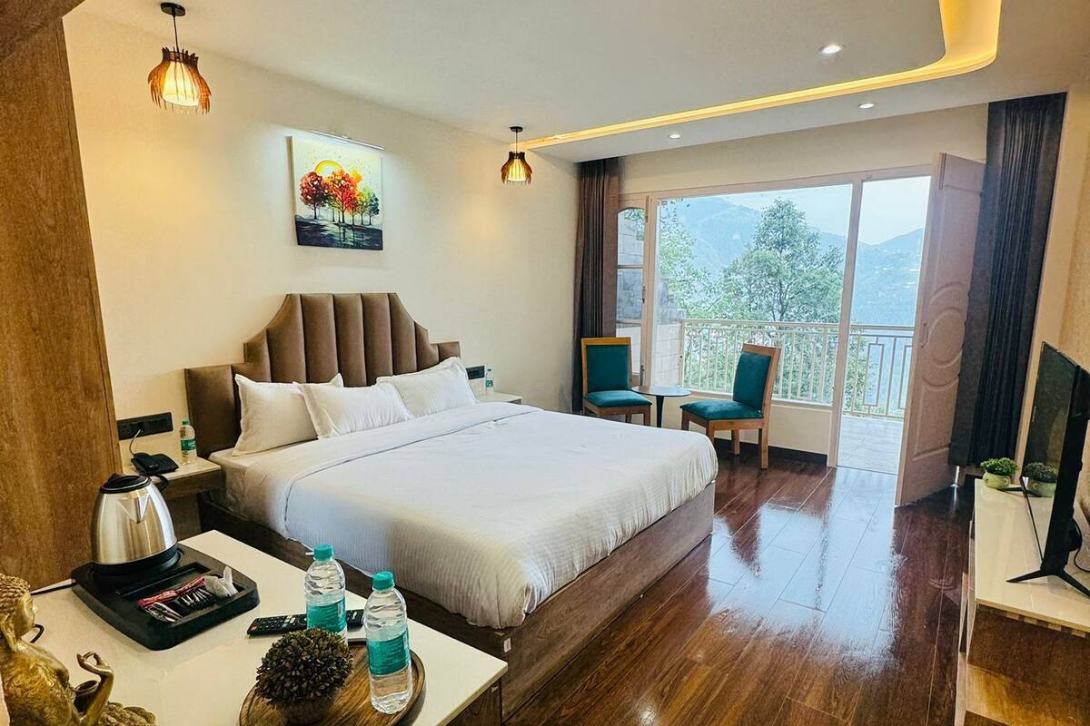Habitación de hotel con una cama grande, un escritorio con hervidor, botellas de agua y un adorno. Hay un cuadro en la pared y vistas al exterior a través de un ventanal amplio.