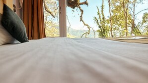 Una cama con sábanas blancas, vistas a árboles y montañas a través de una ventana, y una cortina marrón.
