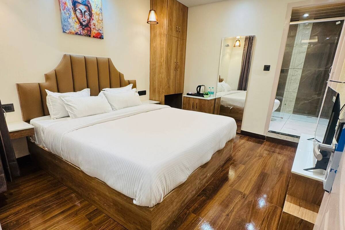 Una habitación de hotel moderna con una cama grande, cabecera de madera, un cuadro en la pared, una mesita con un hervidor y un baño con ducha de vidrio.