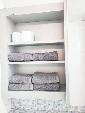 A bathroom with a white tiled floor, a white shelf with towels, and a white toilet.