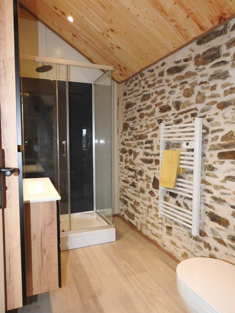 A modern bathroom with a glass shower enclosure, a wooden vanity, a stone wall, and a white towel rack.