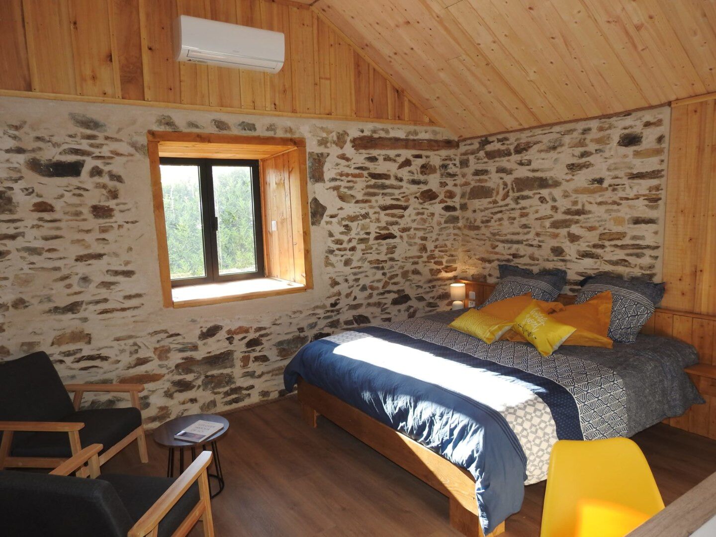 A cozy bedroom with a stone wall, a wooden ceiling, a bed with patterned bedding, a small table, and a chair.