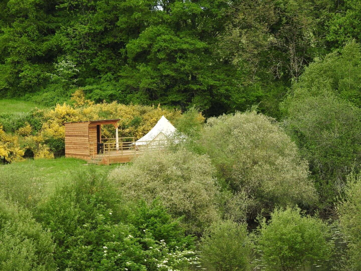 Uma cabana de madeira com deck cercado, rodeada por vegetação densa e uma estrutura em forma de tenda branca.