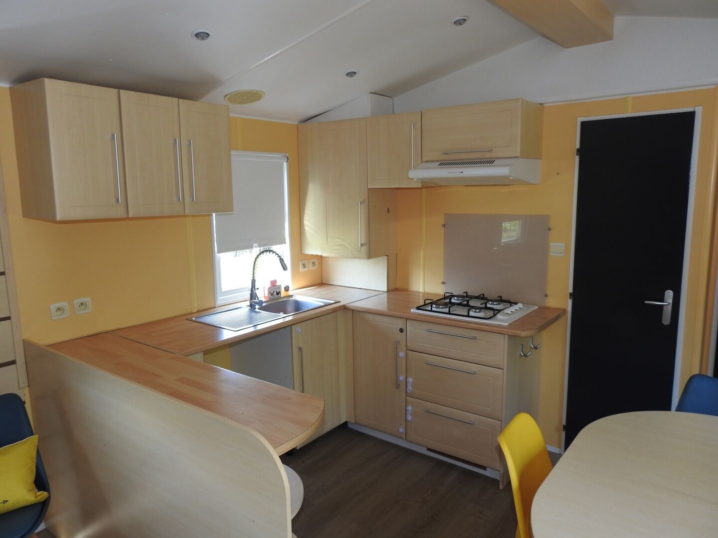 A compact kitchen with wooden cabinets, a gas stove, a sink, and a window. There are two chairs and a table in the dining area.
