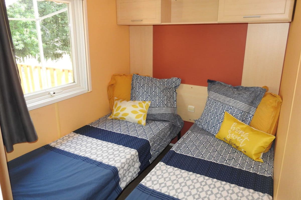 A compact bedroom with two beds, each with patterned bedding and pillows, a window with a view of greenery, and a wooden cabinet above the beds.