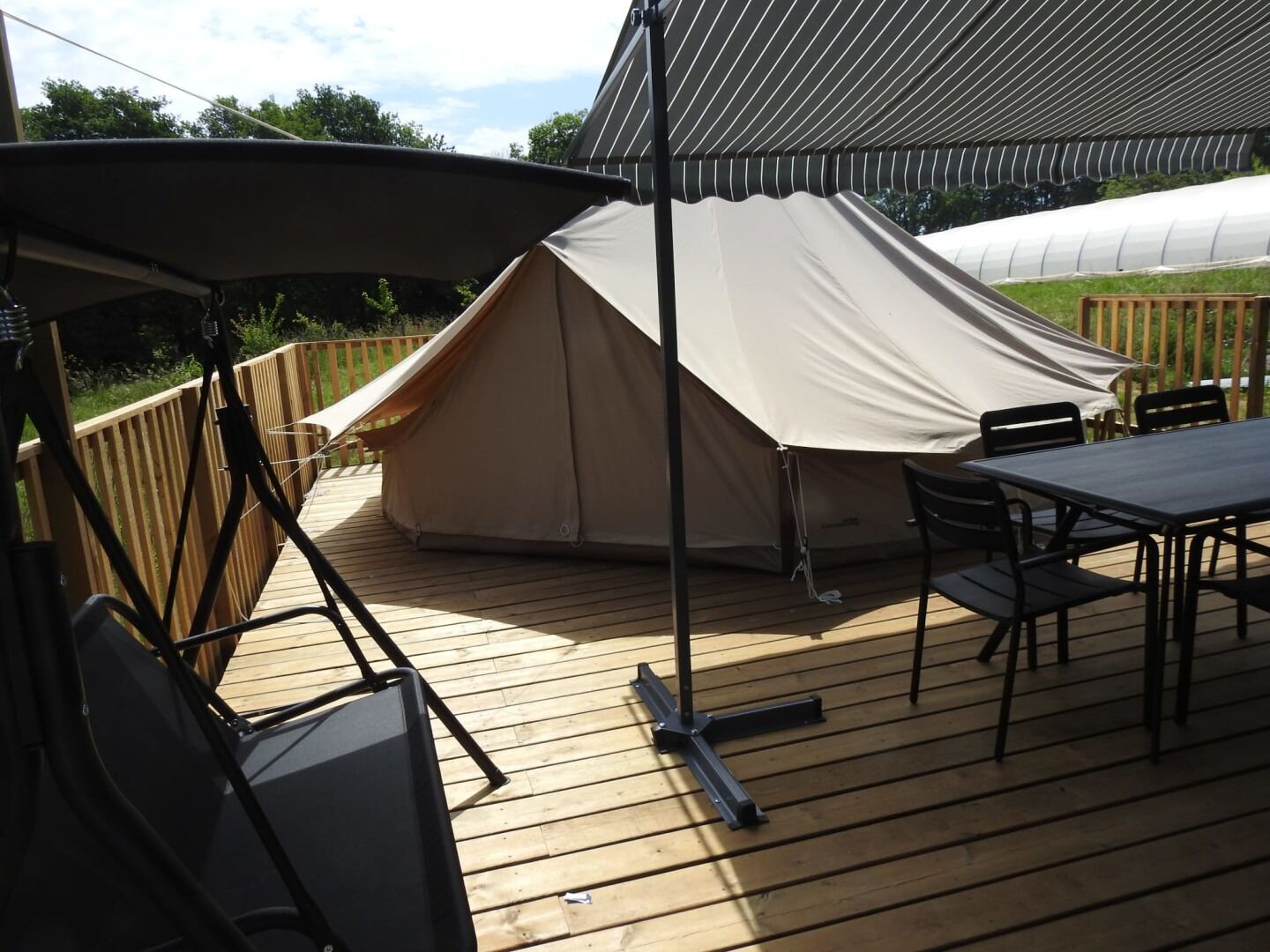Un espace extérieur aménagé avec une tente, une table et des chaises, une terrasse en bois et, au fond, une structure ressemblant à une serre.