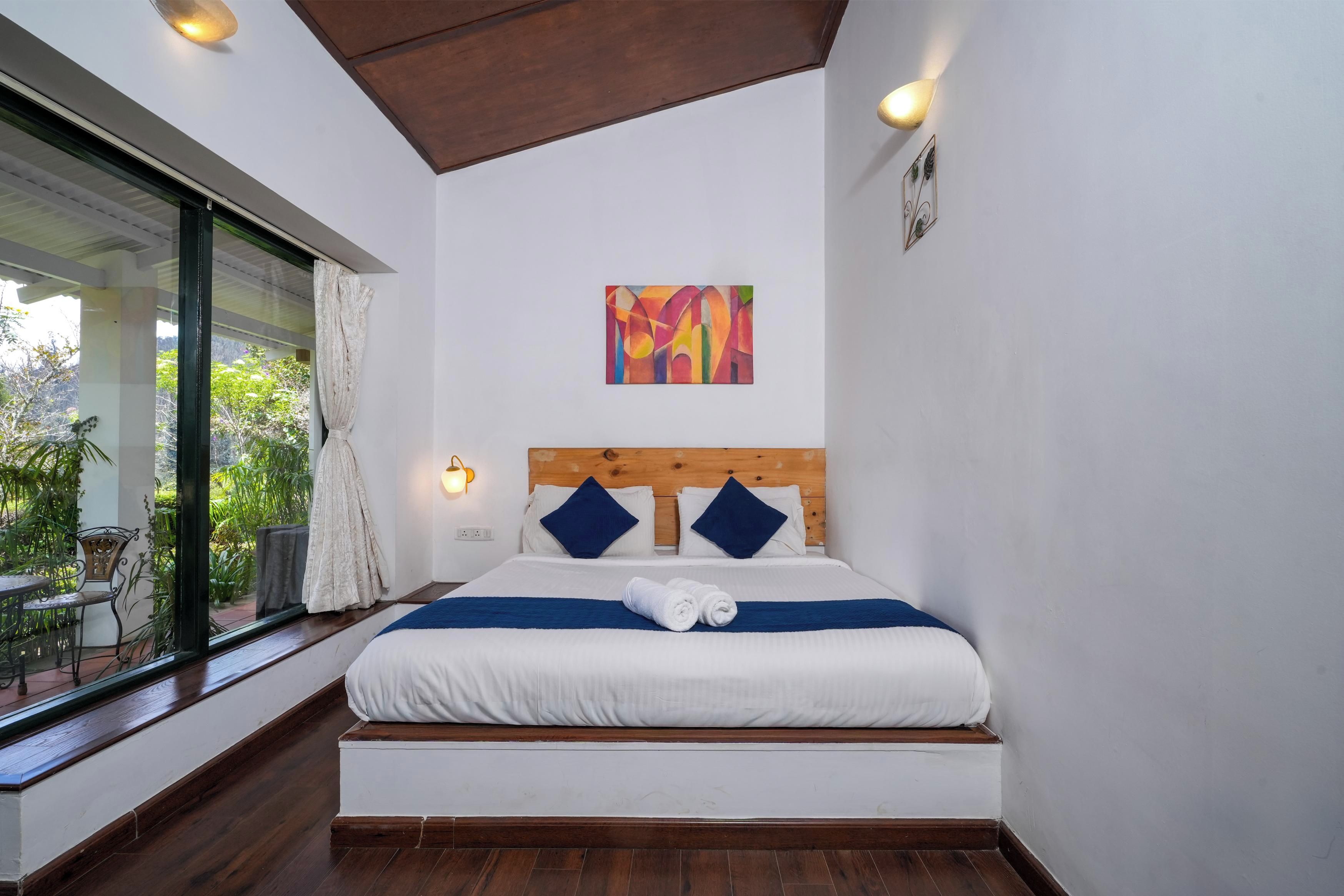 Ein Hotelzimmer mit großem Fenster, einem Bett mit weißer Bettwäsche und blauen Kissen, einem Holz-Kopfschild und einem farbenfrohen, abstrakten Gemälde an der Wand.