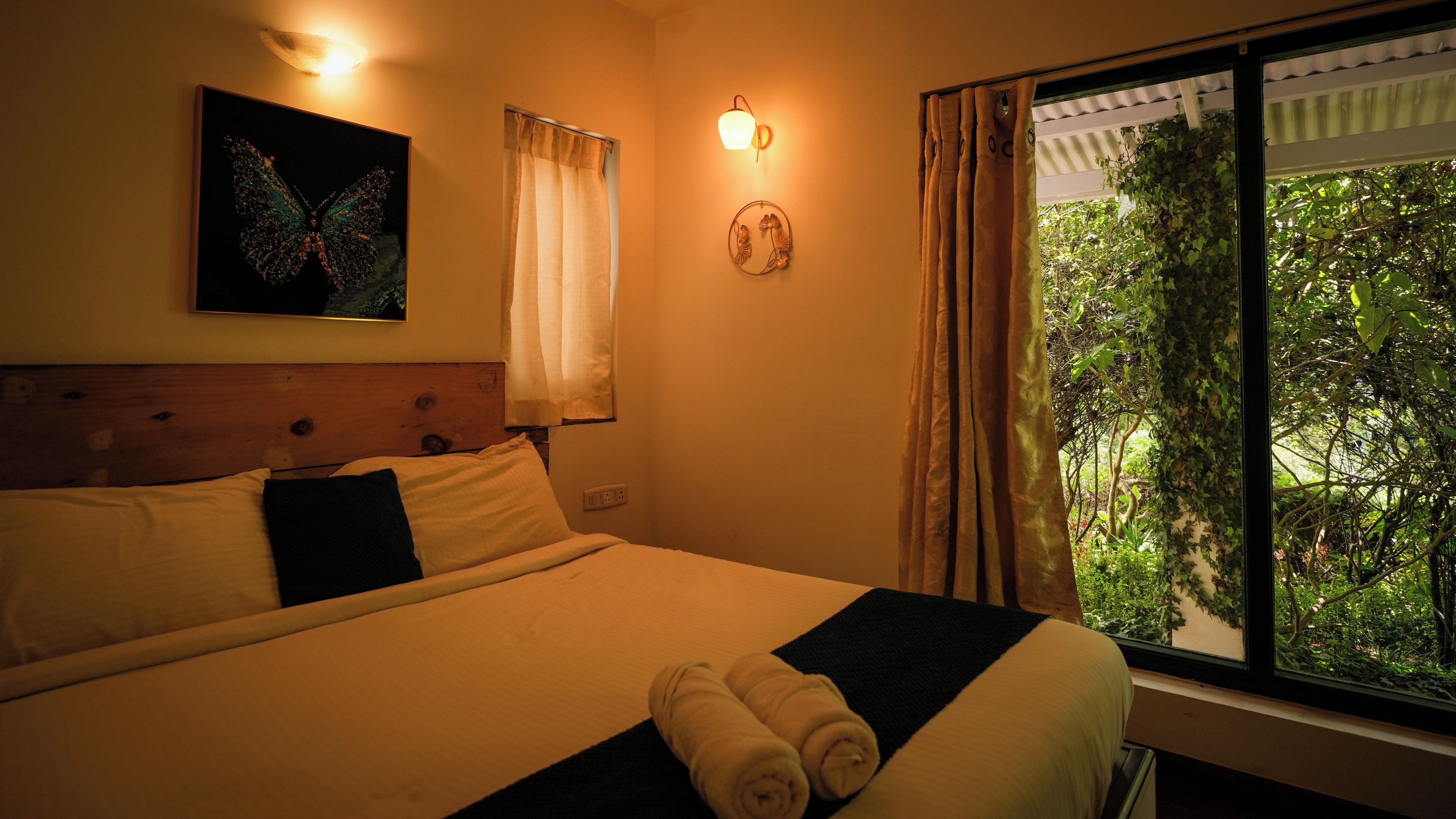 Habitación de hotel con cama, luz empotrada en la pared, ventana con cortinas y un cuadro de una mariposa.
