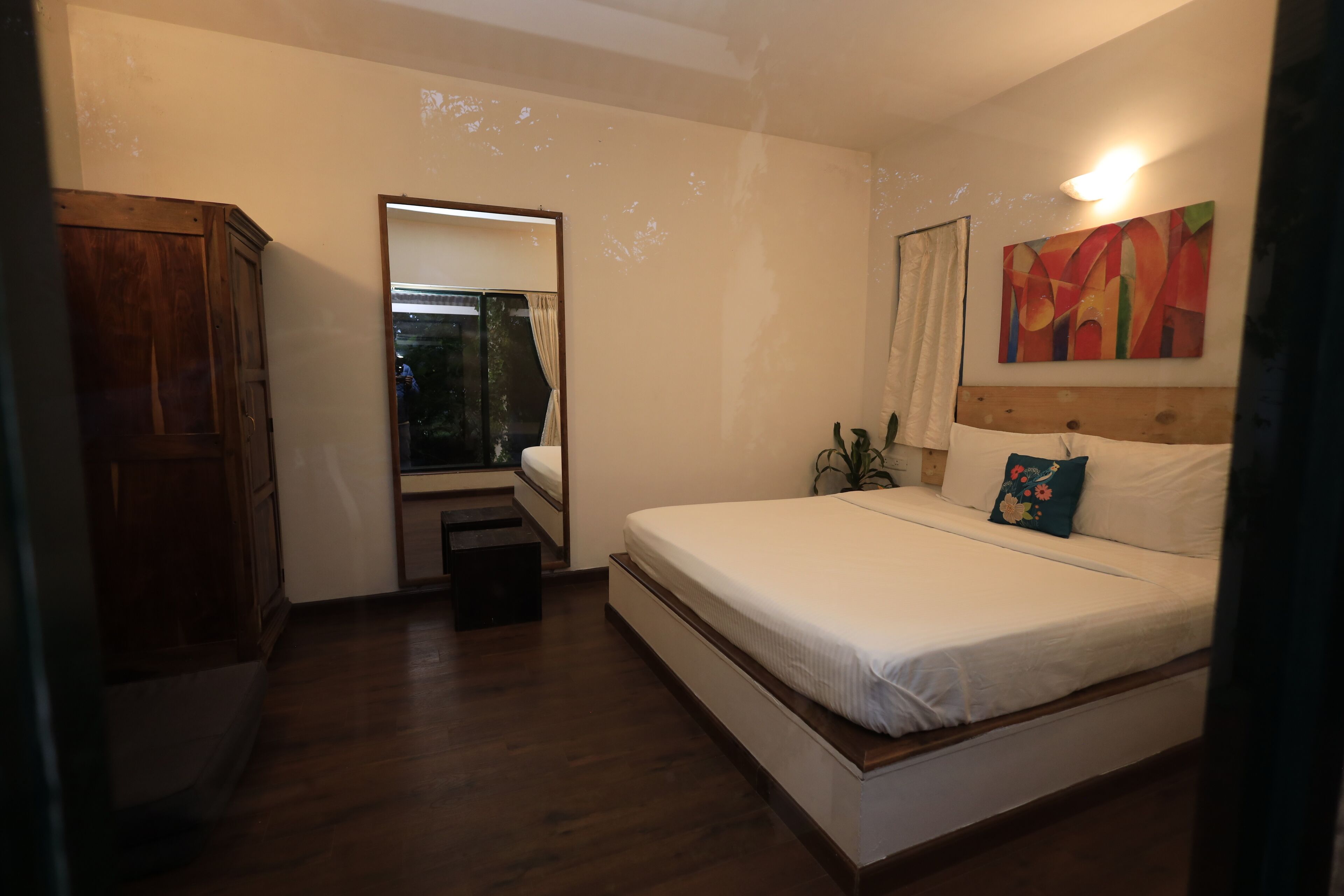 A bedroom with a bed, a wooden wardrobe, a mirror, and a painting on the wall.