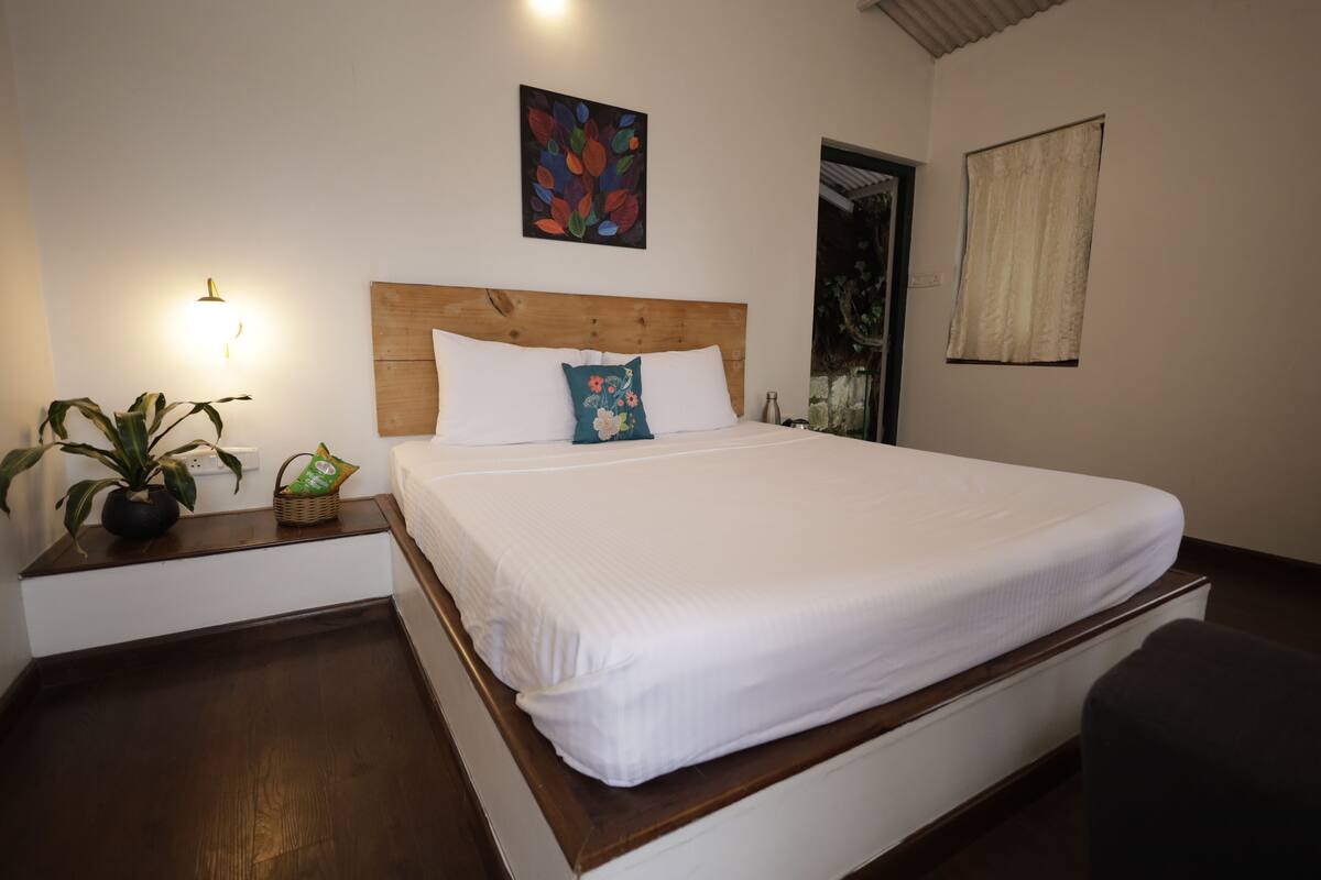 A neatly made bed with white linens, a wooden headboard, and two decorative pillows. A small table with a potted plant and a basket is beside the bed.